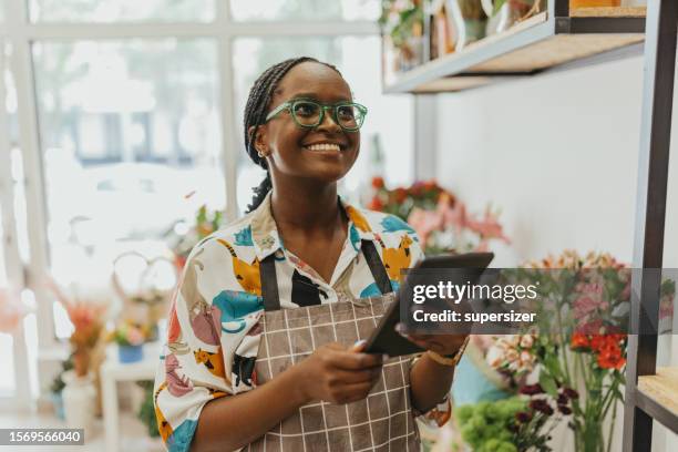 una empresaria afroamericana de 27 años es dueña de una floristería fresca, utilizando una tableta digital. - pequeña empresa fotografías e imágenes de stock