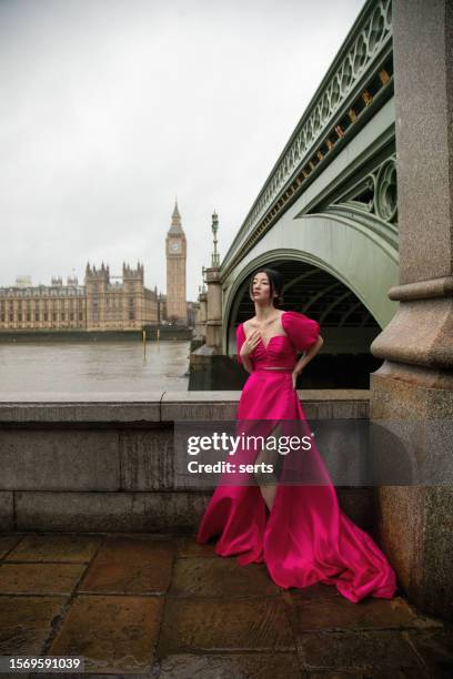 elegante modelo posando con vestido rosa en el icónico puente de westminster con el big ben y el edificio del parlamento de fondo en un día ventoso y lluvioso - london fashion fotografías e imágenes de stock