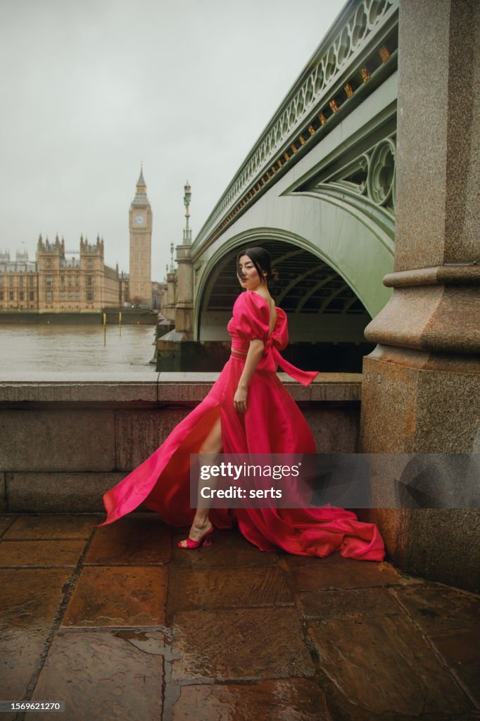 Stylish Fashion Model Posing in Pink Dress at Iconic Westminster Bridge with Big Ben and Parliament Building in Background on a Windy, Rainy Day