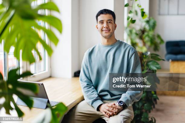 portrait of smiling man sitting by window using laptop - endast en ung man bildbanksfoton och bilder