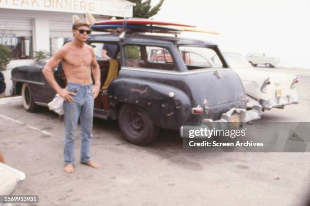 Jan Michael Vincent barechested standing beside car in a scene from the 1978 surfing movie 'Big Wednesday'.