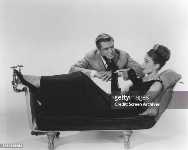 Audrey Hepburn relaxes on couch in elegant dress as Holly Golightly as George Peppard stands behind in a publicity portrait from 'Breakfast at...