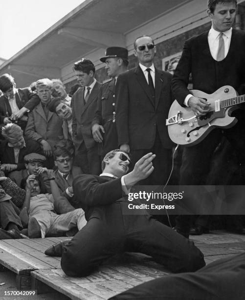 Man in a black suit, kneeling on the dock, accompanied by a guitarist at his side, with a small crowd in the background, during a performance in...