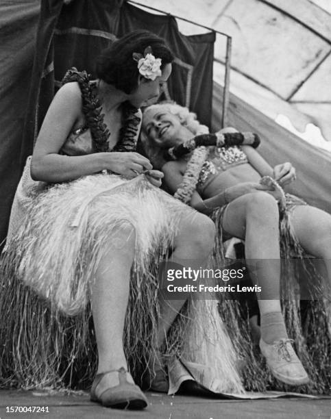 Two women wearing grass skirts and floral garlands, one with a flower in her hair, as the other leans back smiling, circa 1945.