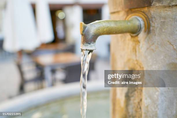 water flowing from a rusty faucet in an outdoor fountain, in the background are some chairs, a table and a parasol. - fuente estructura creada por el hombre fotografías e imágenes de stock