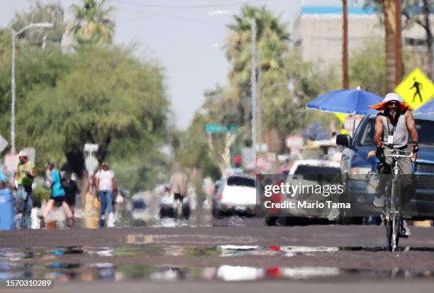 Person rides a bicycle as heat waves shimmer, causing visual distortion, as people walk in the 'The Zone', Phoenix's largest homeless encampment,...