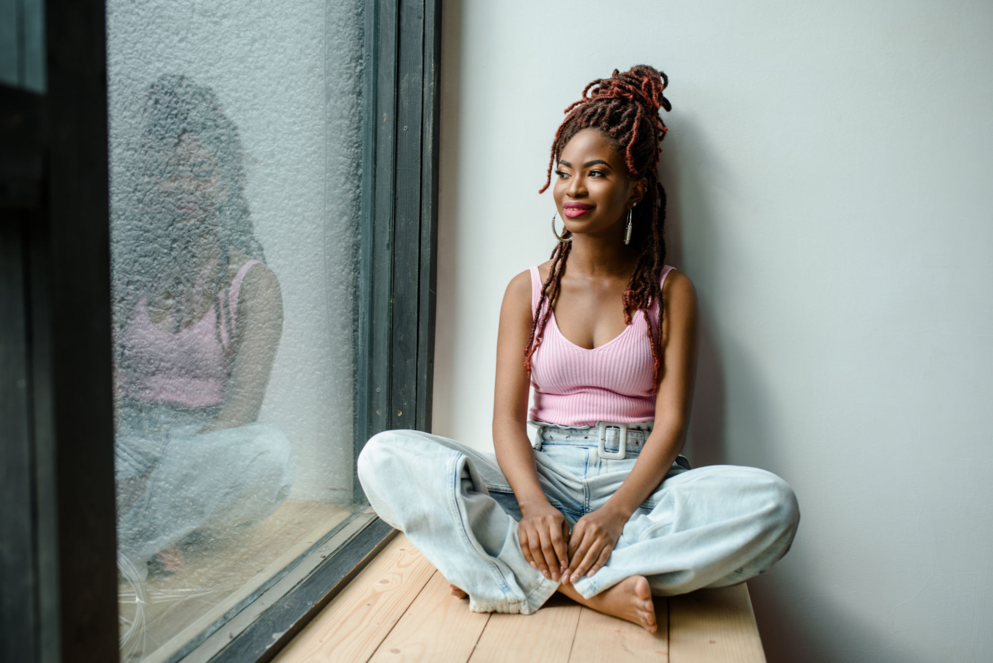 Attractive smiling African woman wearing ribbed tank top and jeans sitting cross-legged on the windowsill and looking through the window. Urban life concept, daydreaming. Planning future concept Attractive smiling African woman wearing ribbed tank top and jeans sitting cross-legged on the windowsill and looking through the window. Urban life concept, daydreaming. Planning future concept