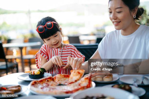 joyful young asian mother and lovely little daughter sharing pizza for lunch in an outdoor restaurant. family enjoying bonding time and a happy meal together. family and eating out lifestyle - pizza restaurant stock pictures, royalty-free photos & images