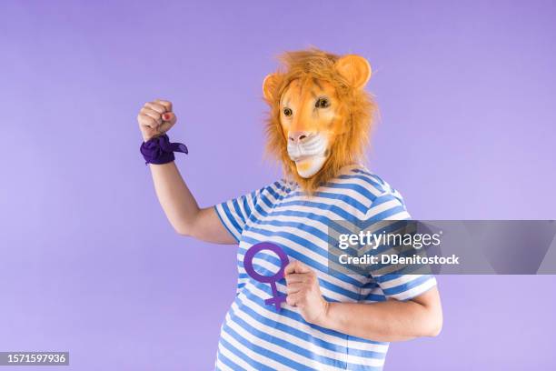 woman wearing blue striped t-shirt and lion mask, holding woman symbol and clenching her fist, on purple background. concept of costume, animal, carnival, feminism, women's rights, party and bizarre. - cat face mask stock pictures, royalty-free photos & images