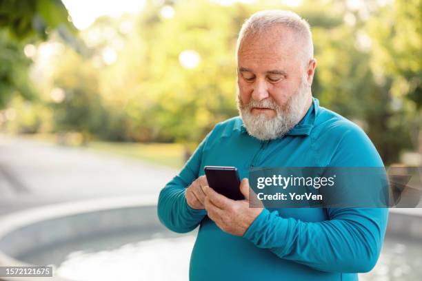 hombre obeso barbudo usando teléfono móvil en un parque público - obesidad fotografías e imágenes de stock