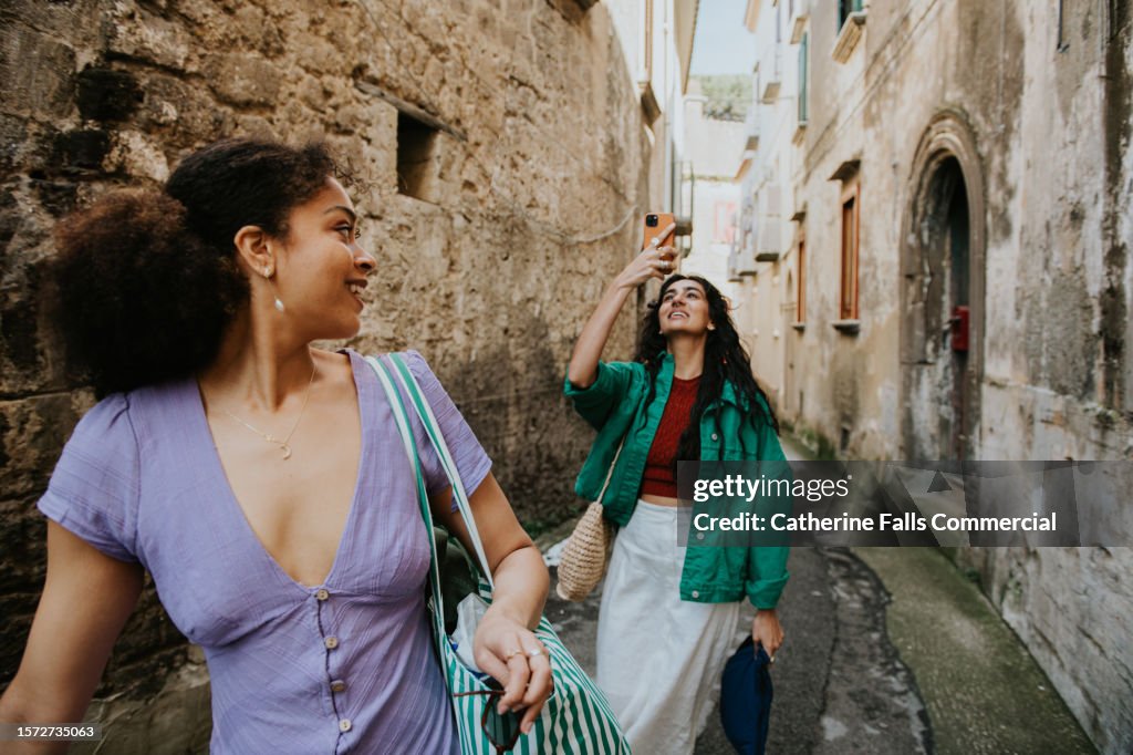Two stylish woman walk down an alleyway. One takes a photo using her mobile phone.