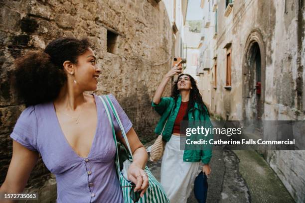 two stylish woman walk down an alleyway. one takes a photo using her mobile phone. - famous women in history stock pictures, royalty-free photos & images