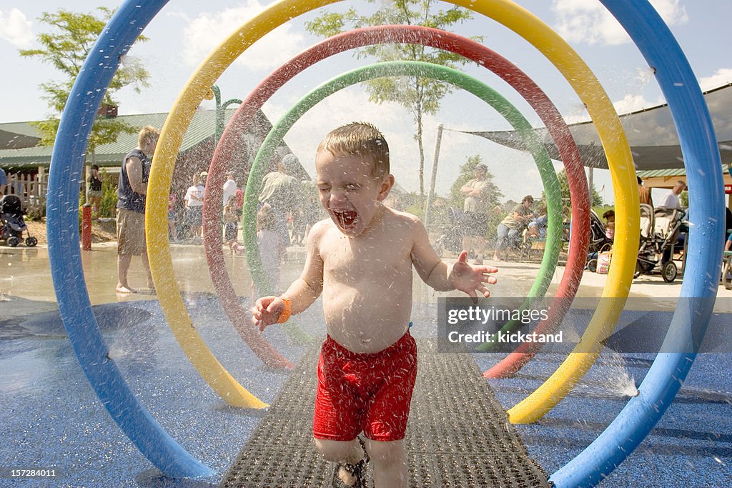 Young laughing boy runs through water park sprinklers