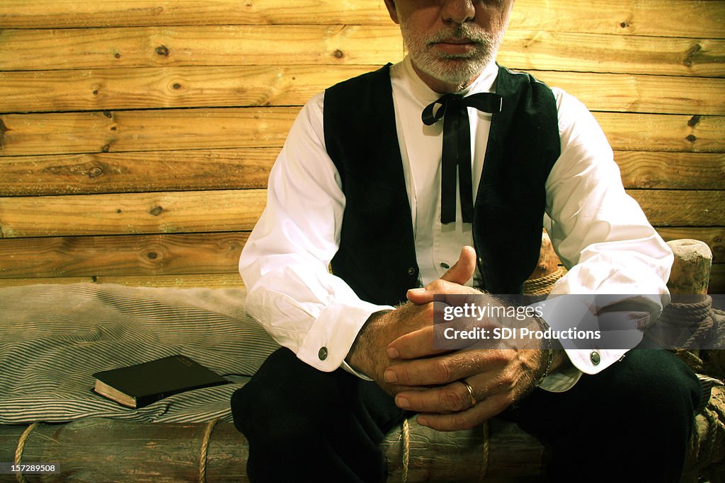 Religious man in a wooden cabin sat on a bed praying