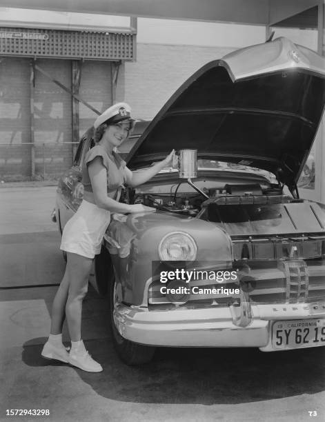 Model in shorts, sneakers and a halter top wearing a Naval cap stands next to an automobile with a raised hood topping up the oil.