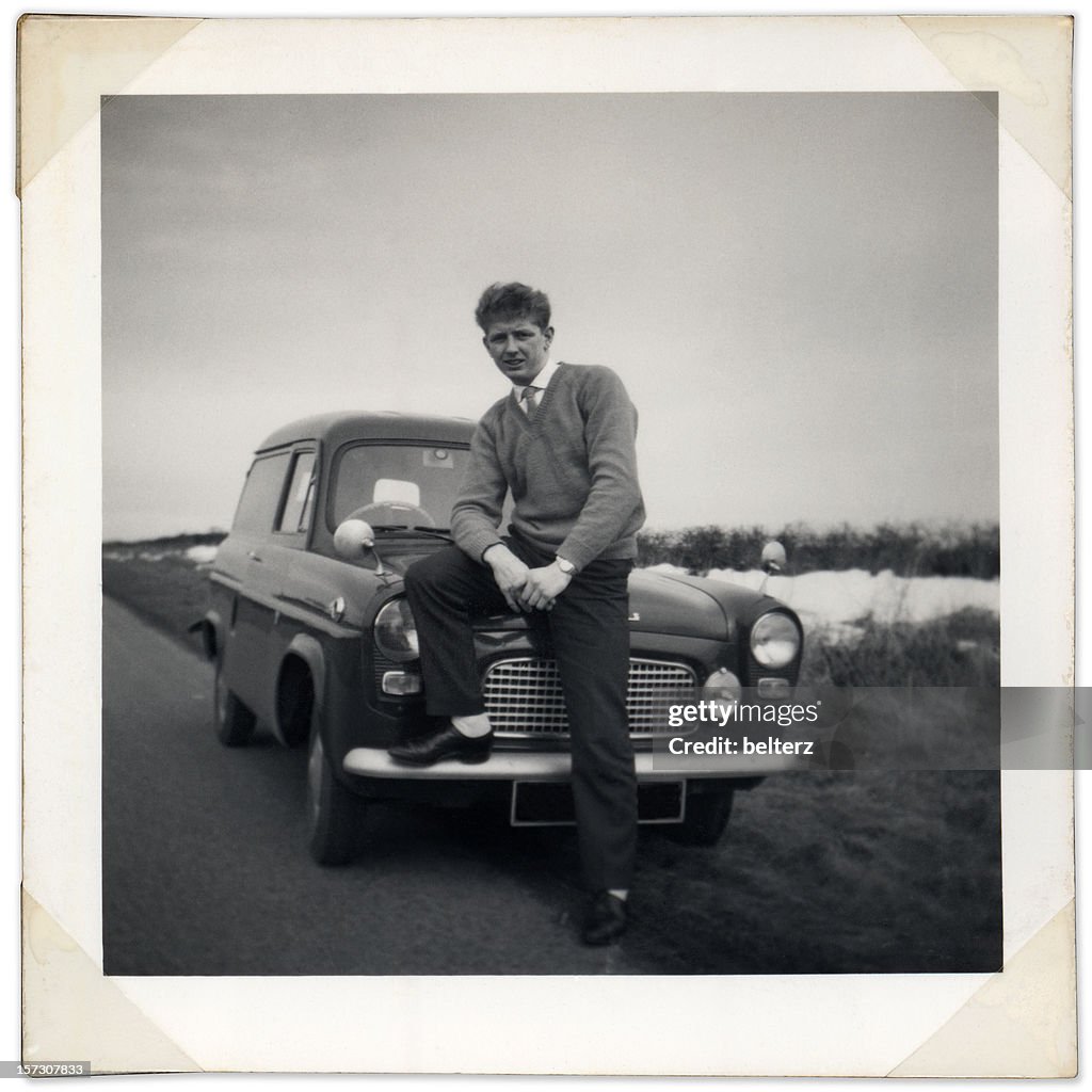 Black and white photo of man sitting on vintage car bonnet