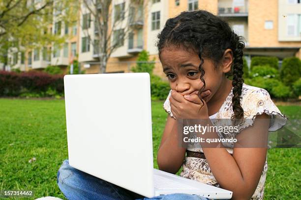 petite fille afro-américaine à l'aide d'ordinateur portable à l'extérieur, révoltés, devant la bouche - fille lire gazon photos et images de collection