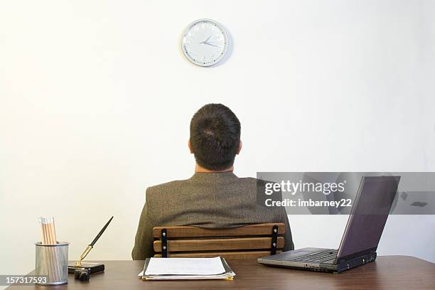 a man sitting backing against a desk looking up at a clock - tijd verspillen stockfoto's en -beelden