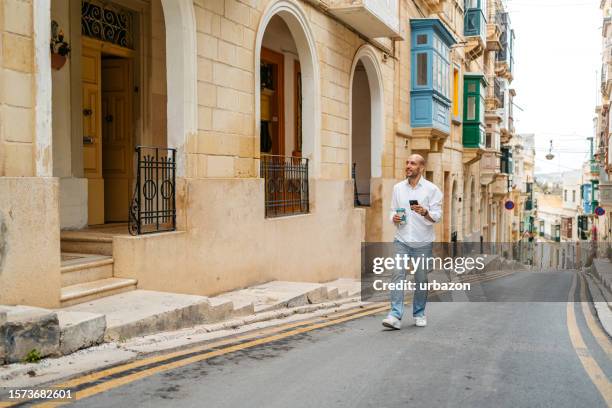 young man drinking coffee and using phone while walking in cospicua malta - malta business stock pictures, royalty-free photos & images