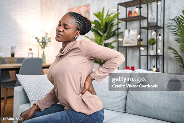 black woman suffering with back pain while sitting at home - back pain stock pictures, royalty-free photos & images