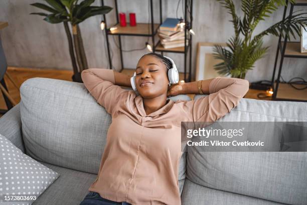black woman listening to music on her wireless headphones and relaxing on her living room sofa - comfort zone bildbanksfoton och bilder