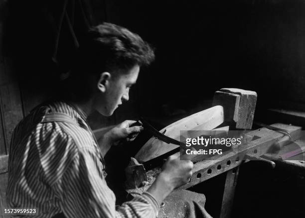 Man uses a hand-held scraper to shape the hull of a toy boat from a block of wood held in a vice, in a workshop, United States, circa 1925.