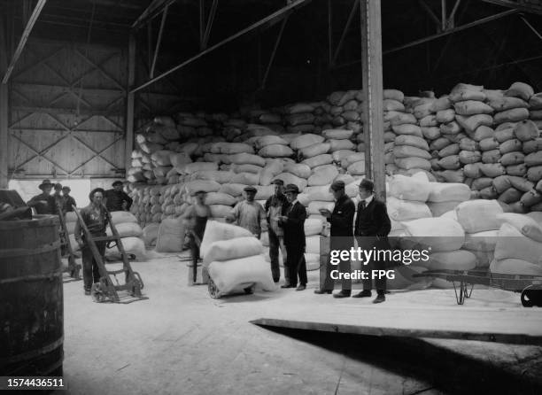 Group of men standing in a warehouse, with sacks piled up in the background, and porters moving sacks around on sack trucks, with a ramp in the...