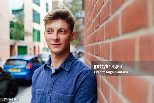 a young man leans against a wall in the city, looking away with a contemplative expression - fashion man single casual shirt stock pictures, royalty-free photos & images