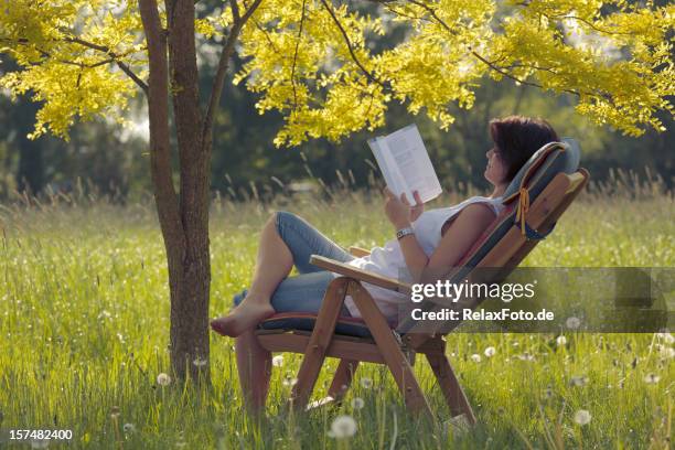 young woman on wooden deck chair under tree reading book - 1 june 2009 stock pictures, royalty-free photos & images
