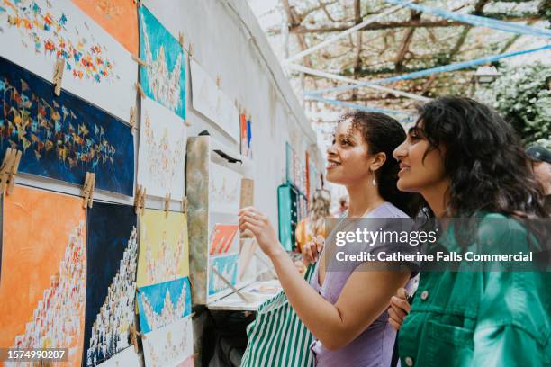 two woman admire paintings at a market stall as they vacation in europe - exhibition fotografías e imágenes de stock