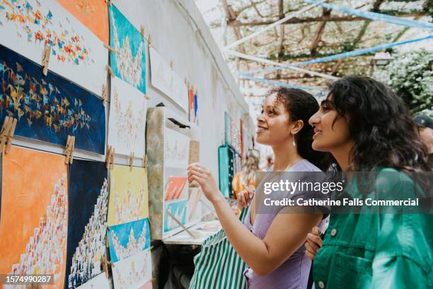 two woman admire paintings at a market stall as they vacation in europe - tentoonstelling stockfoto's en -beelden
