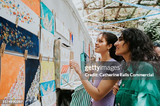 Two woman admire paintings at a market stall as they vacation in Europe