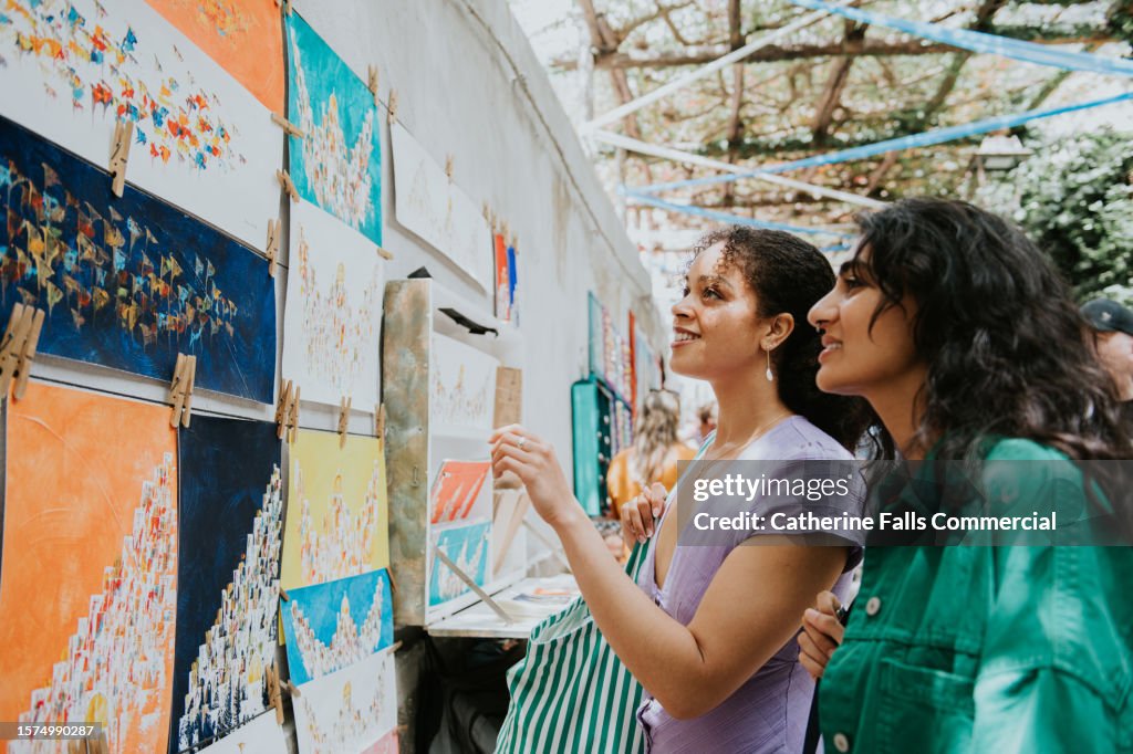 Two woman admire paintings at a market stall as they vacation in Europe