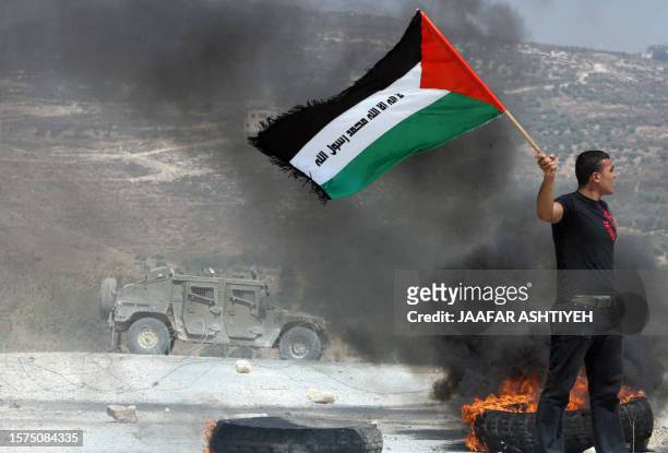 Palestinian demonstrator holds a Palestinian flag next to buring tires as an Israeli military vehicle stands in the background during a protest...