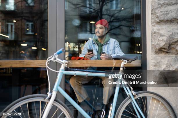 thoughtful male person in warm clothes sits in cafe looking out panoramic window with parked bike - center athlete stock pictures, royalty-free photos & images
