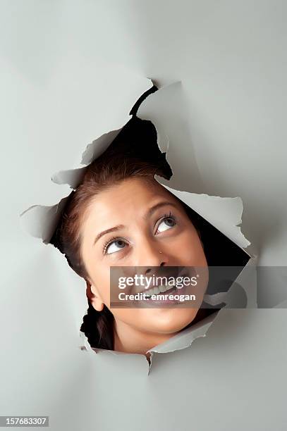 young woman looking out through hole in grey paper - gluren stockfoto's en -beelden
