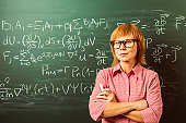 Female teacher in horn rimmed glasses is standing against blackboard