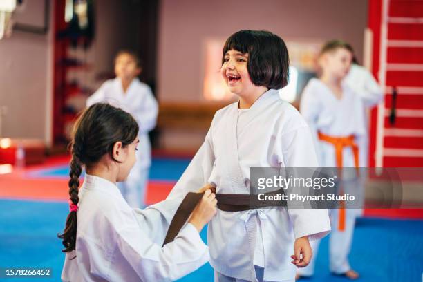 a group of elementary age children are taking a martial arts class. friends tying a belt in a dojo - jujitsu stock pictures, royalty-free photos & images