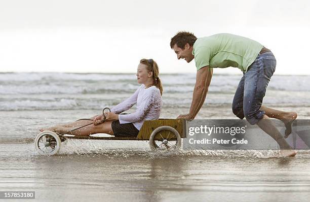 father pushing daughter in go-kart on beach - mann schiebt stock-fotos und bilder