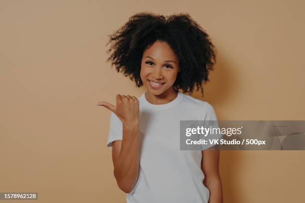 young smiling dark skinned female curly hair pointing aside with thumb,isolated over beige wall - editorial template stock pictures, royalty-free photos & images