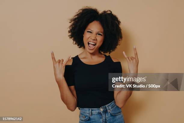 young african woman with hands in rock n roll sign and shouting loudly,isolated on beige background - editorial template stock pictures, royalty-free photos & images