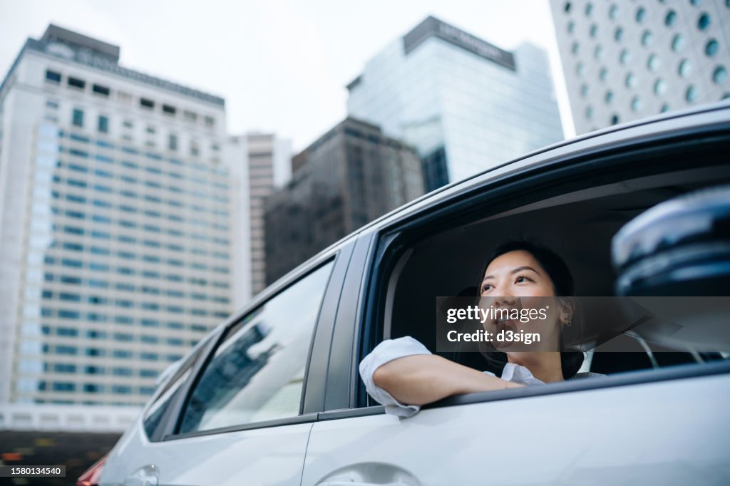 Young Asian woman sitting in backseat of a car, looking out of window and smiling while commuting in the city