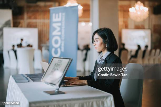 asiatische malaiische rezeptionistin mit laptop am eingang des sitzungssaals, die sich auf die registrierung der teilnehmer vorbereitet - einschreibungsveranstaltung stock-fotos und bilder
