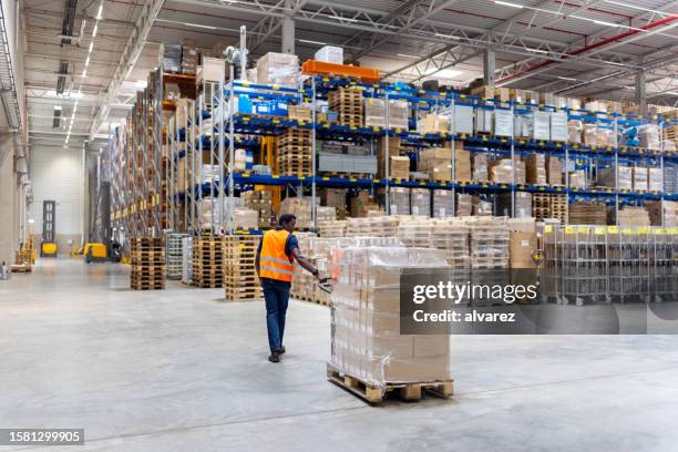 young warehouse worker pulling pallet jack with boxes - sala de armazenamento imagens e fotografias de stock