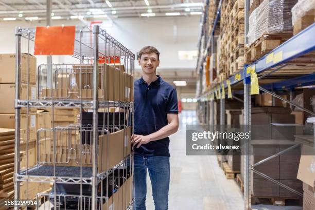portrait of a confident young male worker with trolley in distribution warehouse - distribution warehouse stock pictures, royalty-free photos & images