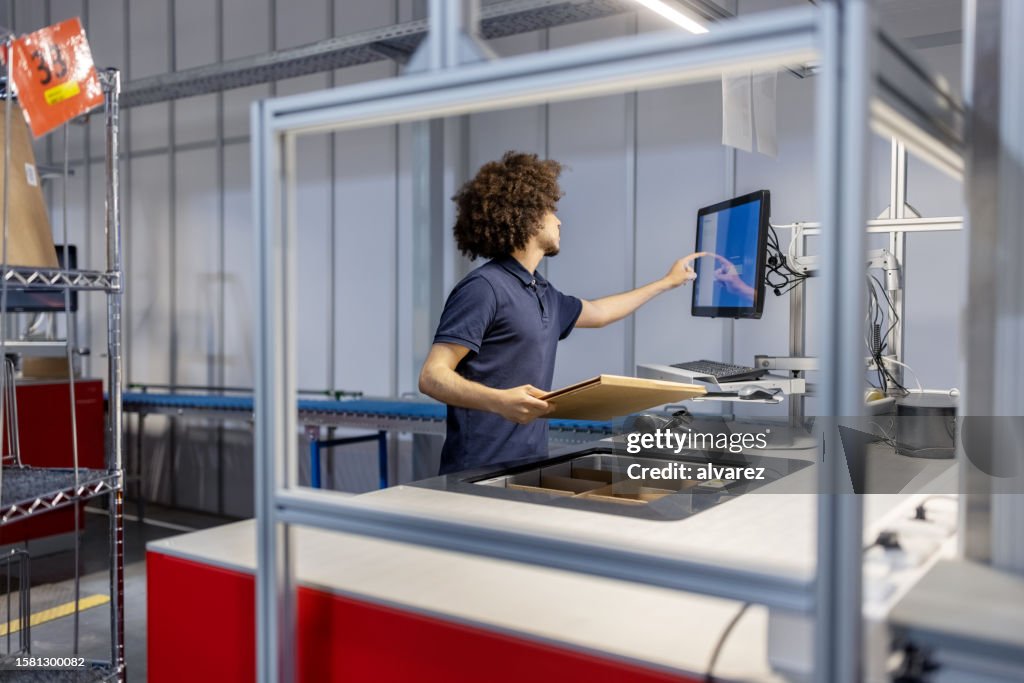 Young man holding a box and updating package details on computer at automated warehouse