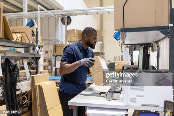 african man holding a parcel and looking at computer screen while working in a warehouse - warehouse packing stock pictures, royalty-free photos & images
