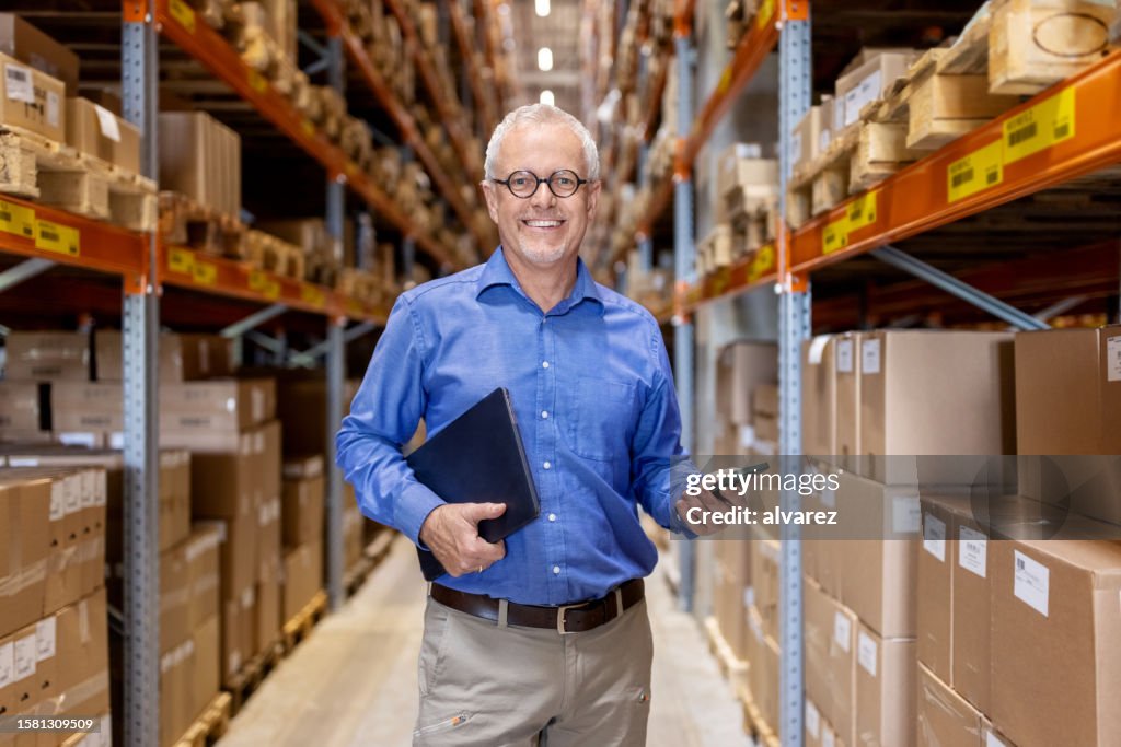 Portrait of smiling warehouse manager standing by racks in storage facility