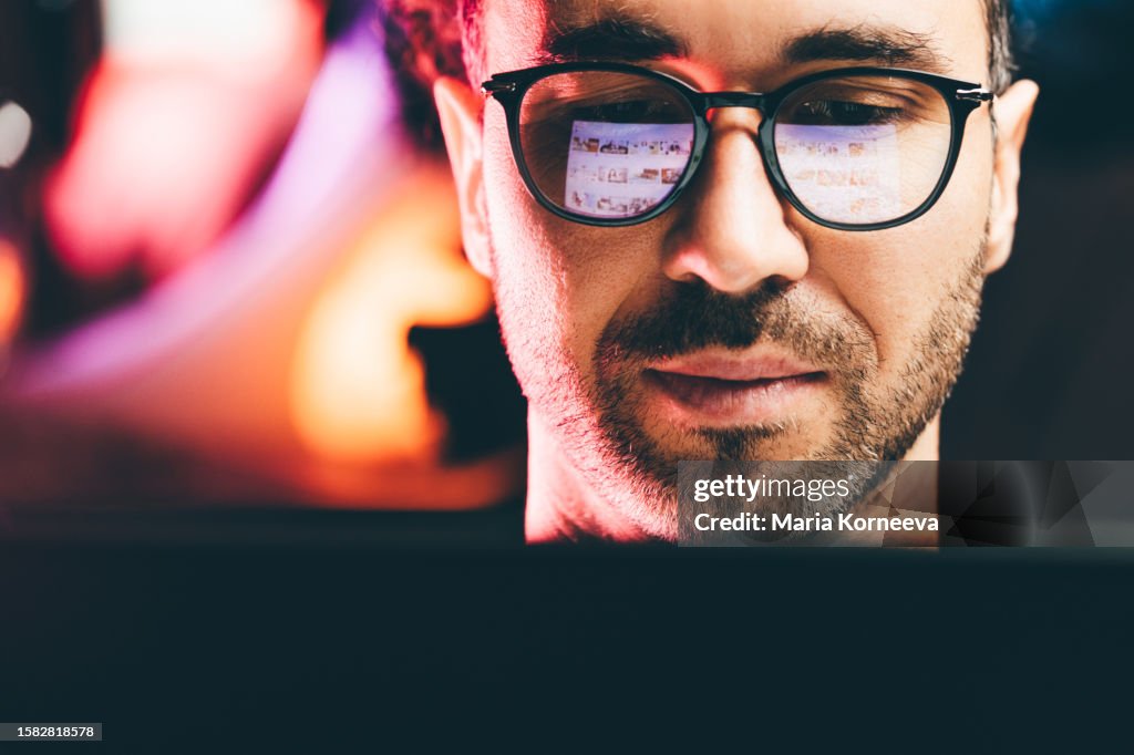Men wearing glasses watching something on his laptop at home at the night. Young man using a laptop, reflection of a website in his glasses, close up.