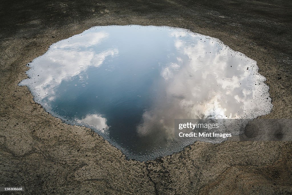 Rain drops falling onto a large puddle. A reflection of sky and clouds.